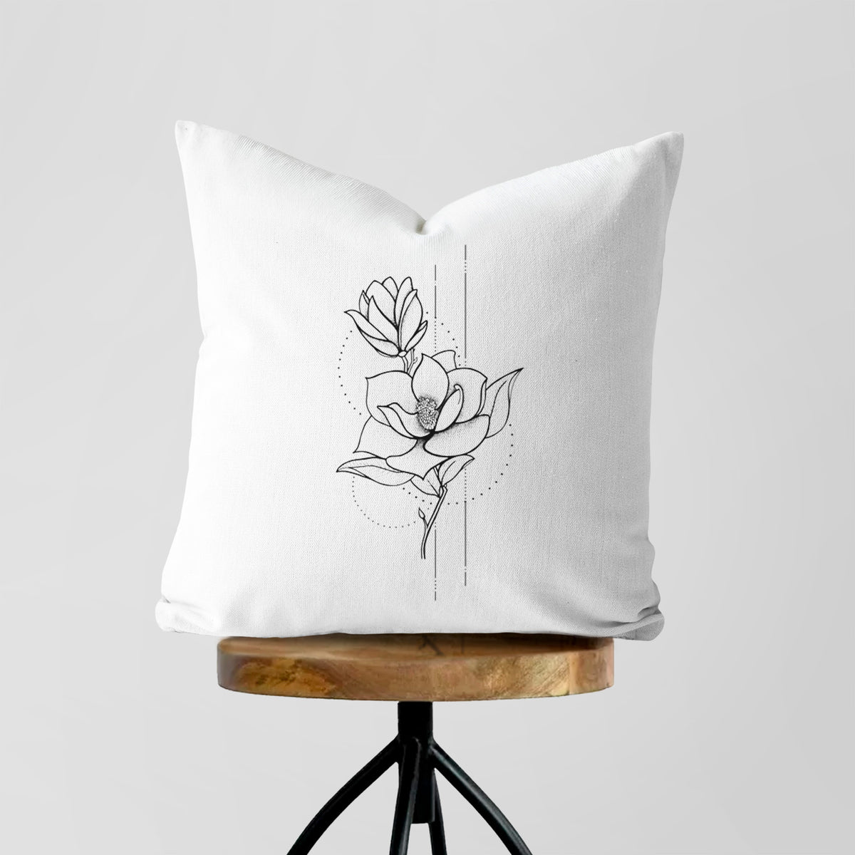 White pillow cover printed with a black ink line drawing of a magnolia blossom on a stem, on a wooden stool.