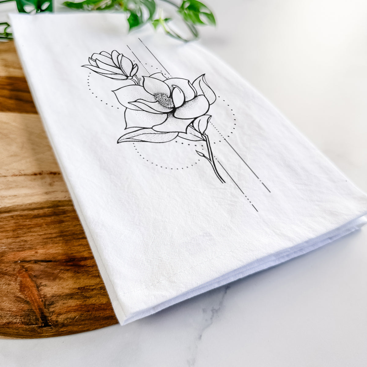 Close-up of a folded white tea towel on a wooden board, showing a black ink line drawing of a magnolia blossom on a stem.