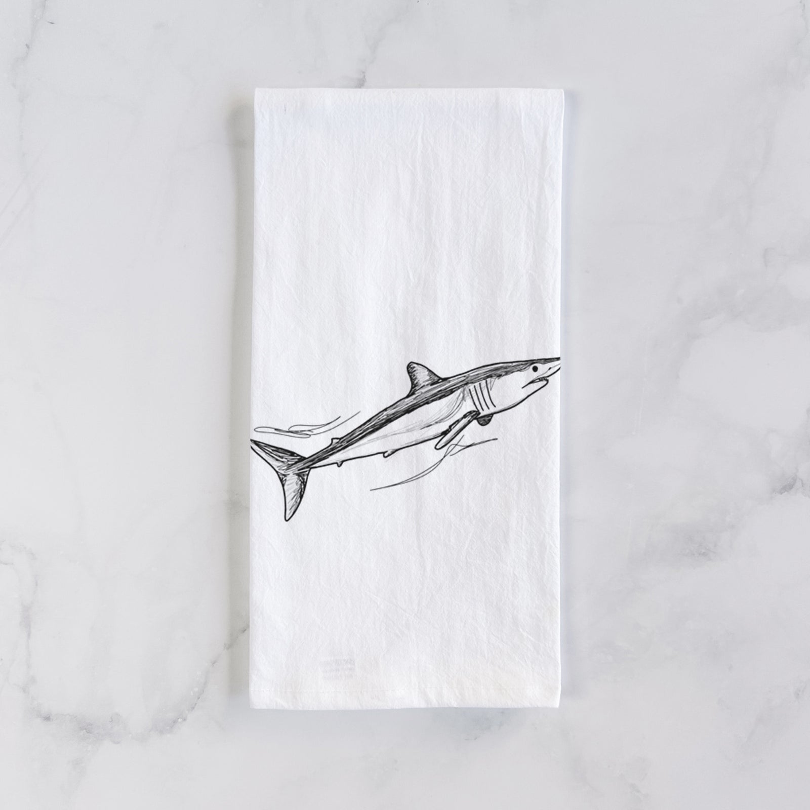 White cotton tea towel printed with a black ink illustration of a mako shark swimming, on a marble counter beside a round wooden cutting board and greenery.
