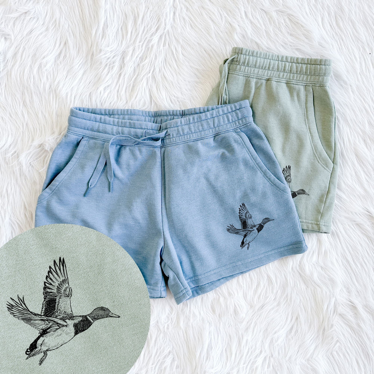 Two pairs of Cali Wave shorts in different colors printed with a detailed black ink illustration of a male mallard duck taking flight, with a close-up of the print.