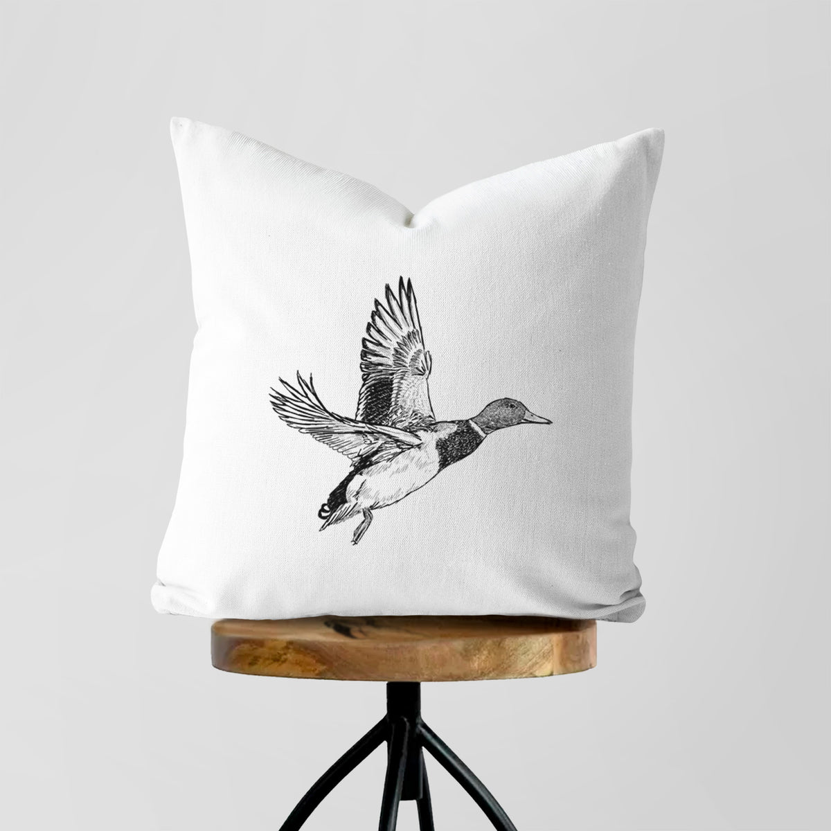 White pillow cover printed with a detailed black ink illustration of a male mallard duck taking flight, on a wooden stool.