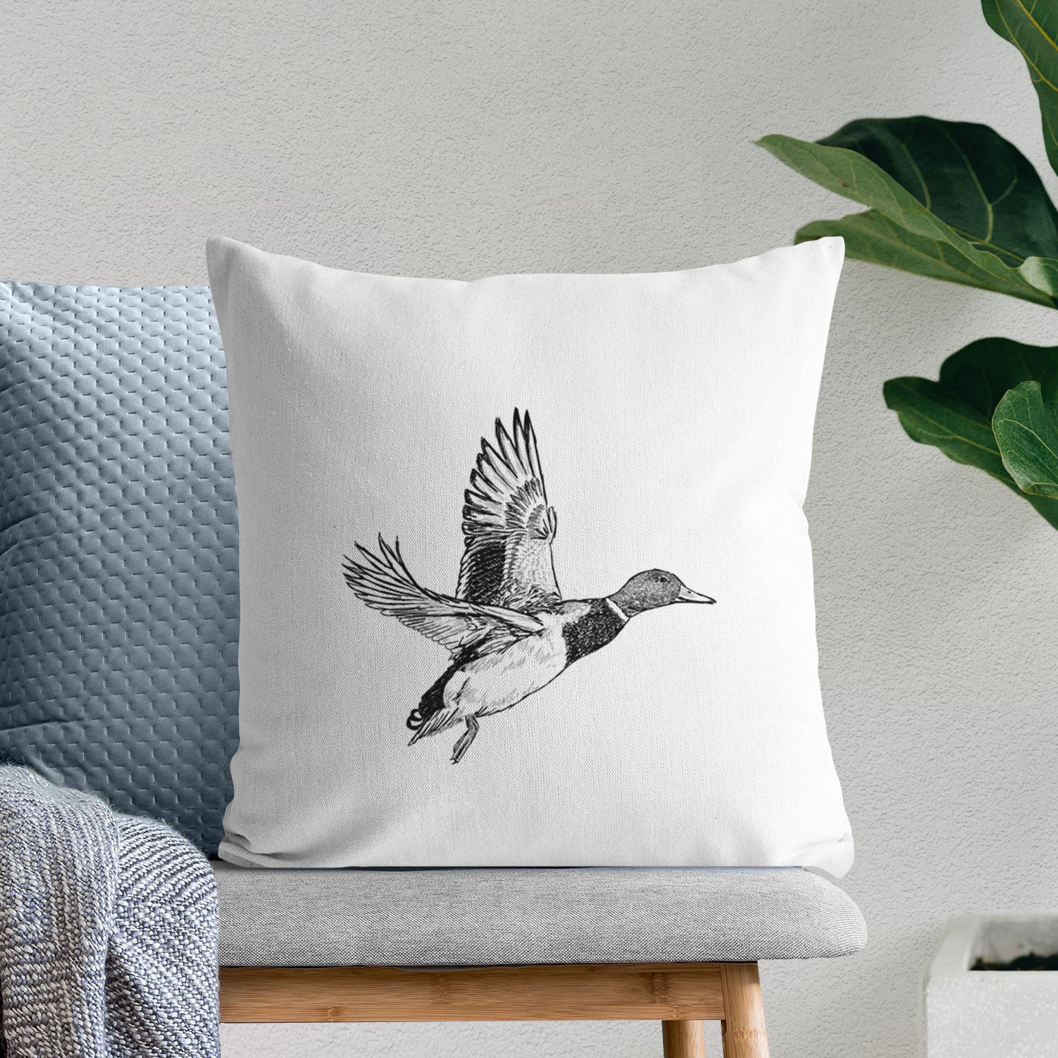 White pillow cover printed with a detailed black ink illustration of a male mallard duck taking flight, on a bench beside plants.