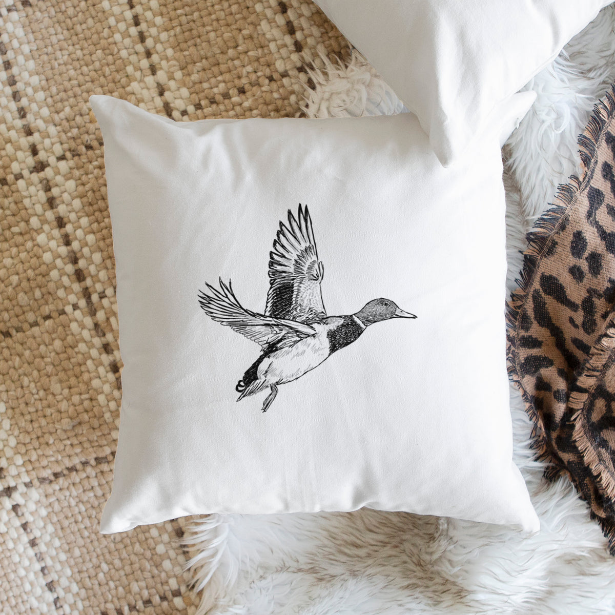White pillow cover printed with a detailed black ink illustration of a male mallard duck taking flight, on a woven chair.