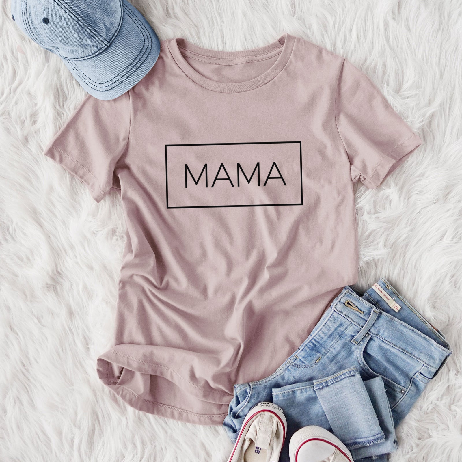 Women's relaxed fit cotton t-shirt printed with the word MAMA in thin black letters inside a rectangle outline, styled flat on a white faux fur rug with jeans.