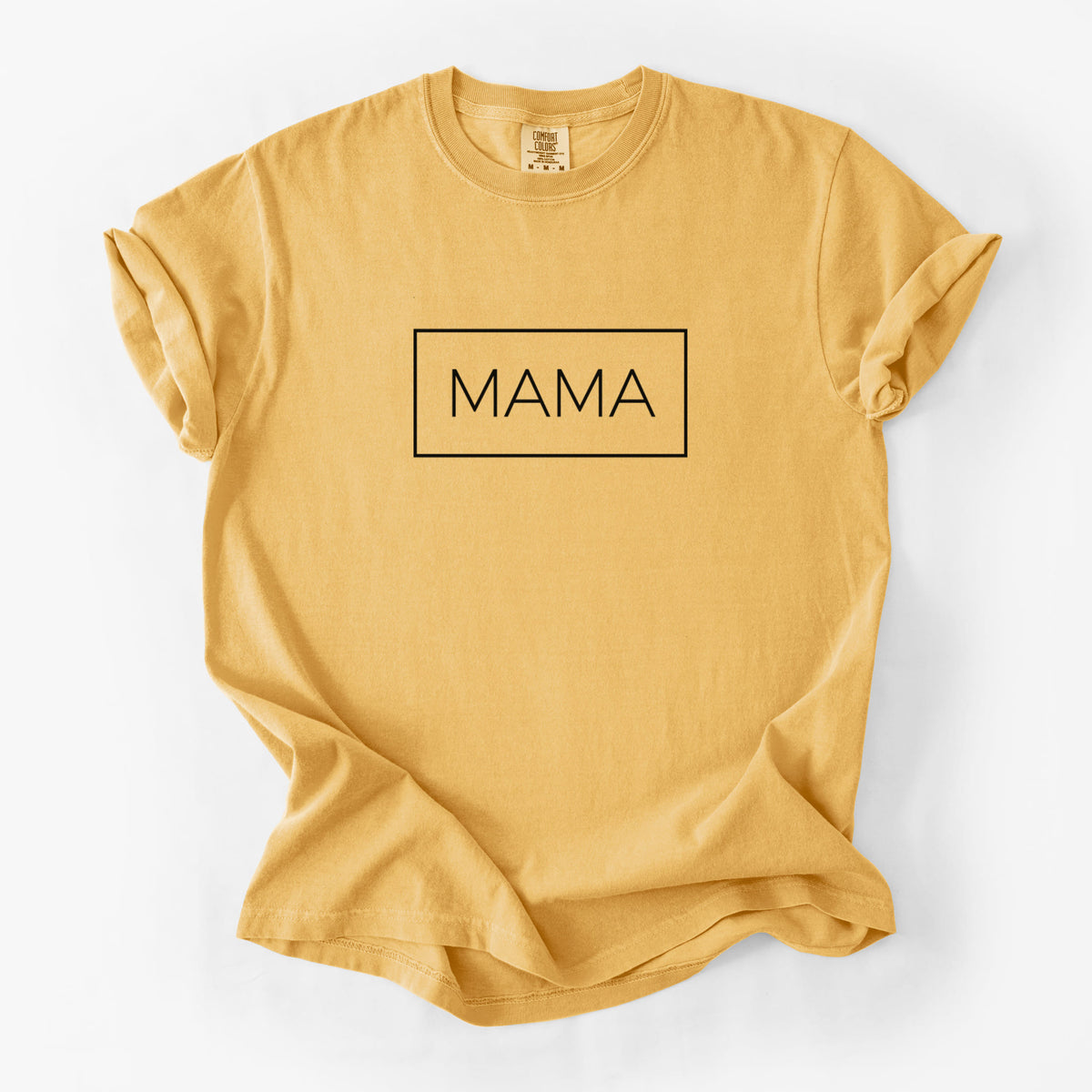 Golden wheat yellow Comfort Colors t-shirt laid flat, printed with the word MAMA in thin black letters inside a rectangle outline.