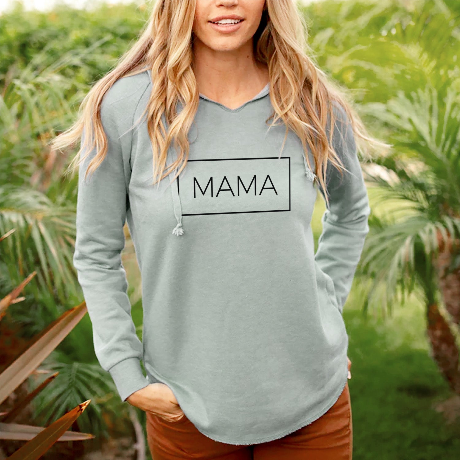 Smiling woman on a seaside bluff wearing a sage green women's Cali Wave hooded sweatshirt printed with the word MAMA in thin black letters inside a rectangle outline.