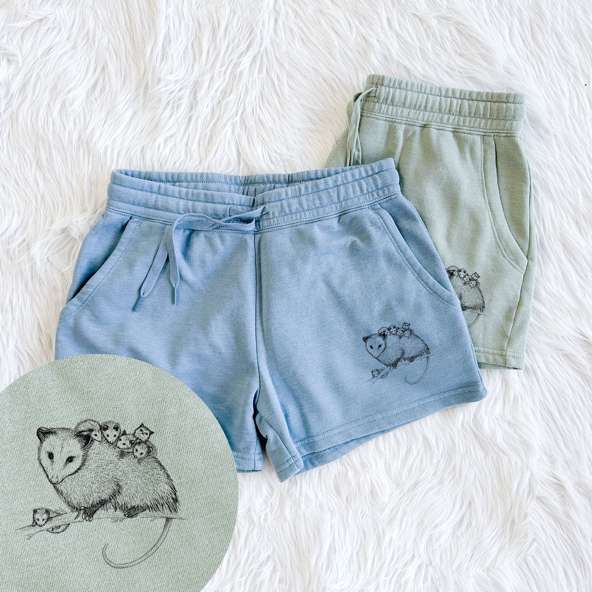 Two pairs of Cali Wave shorts in different colors printed with a black ink illustration of a mother opossum on a branch carrying her babies on her back, with a close-up of the print.