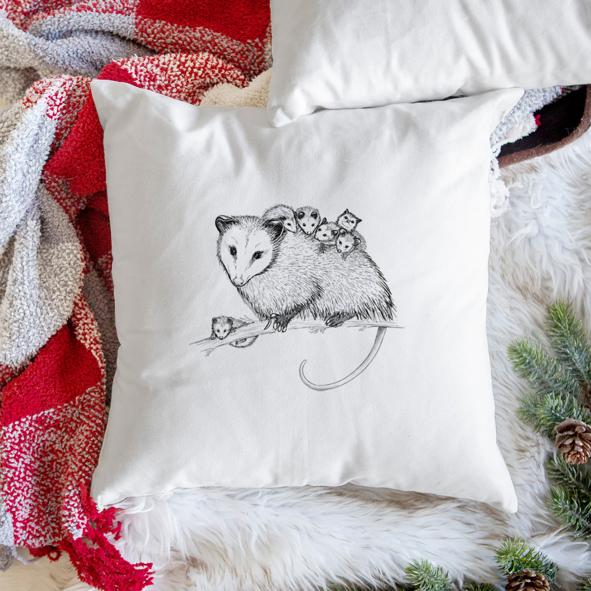 White pillow cover printed with a black ink illustration of a mother opossum on a branch carrying her babies on her back, on a woven chair.