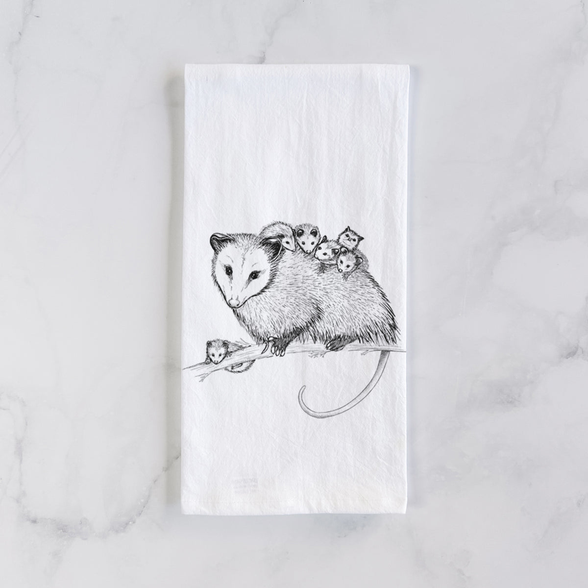 White cotton tea towel laid flat on marble, printed with a black ink illustration of a mother opossum on a branch carrying her babies on her back.