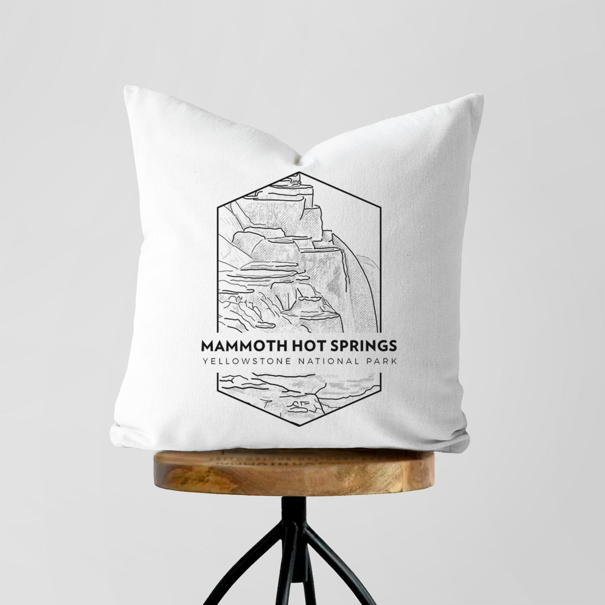 White pillow cover printed with a black line art badge of the terraces at Mammoth Hot Springs, with the words Mammoth Hot Springs, Yellowstone National Park, on a wooden stool.