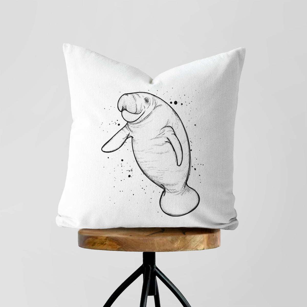 White pillow cover printed with a black ink line drawing of a manatee swimming, with ink splatters, on a wooden stool.