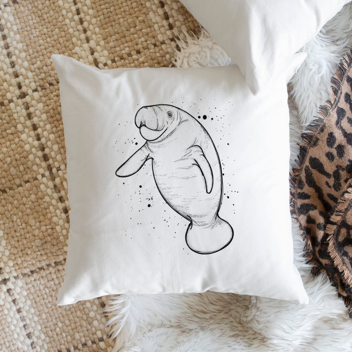 White pillow cover printed with a black ink line drawing of a manatee swimming, with ink splatters, on a woven chair.