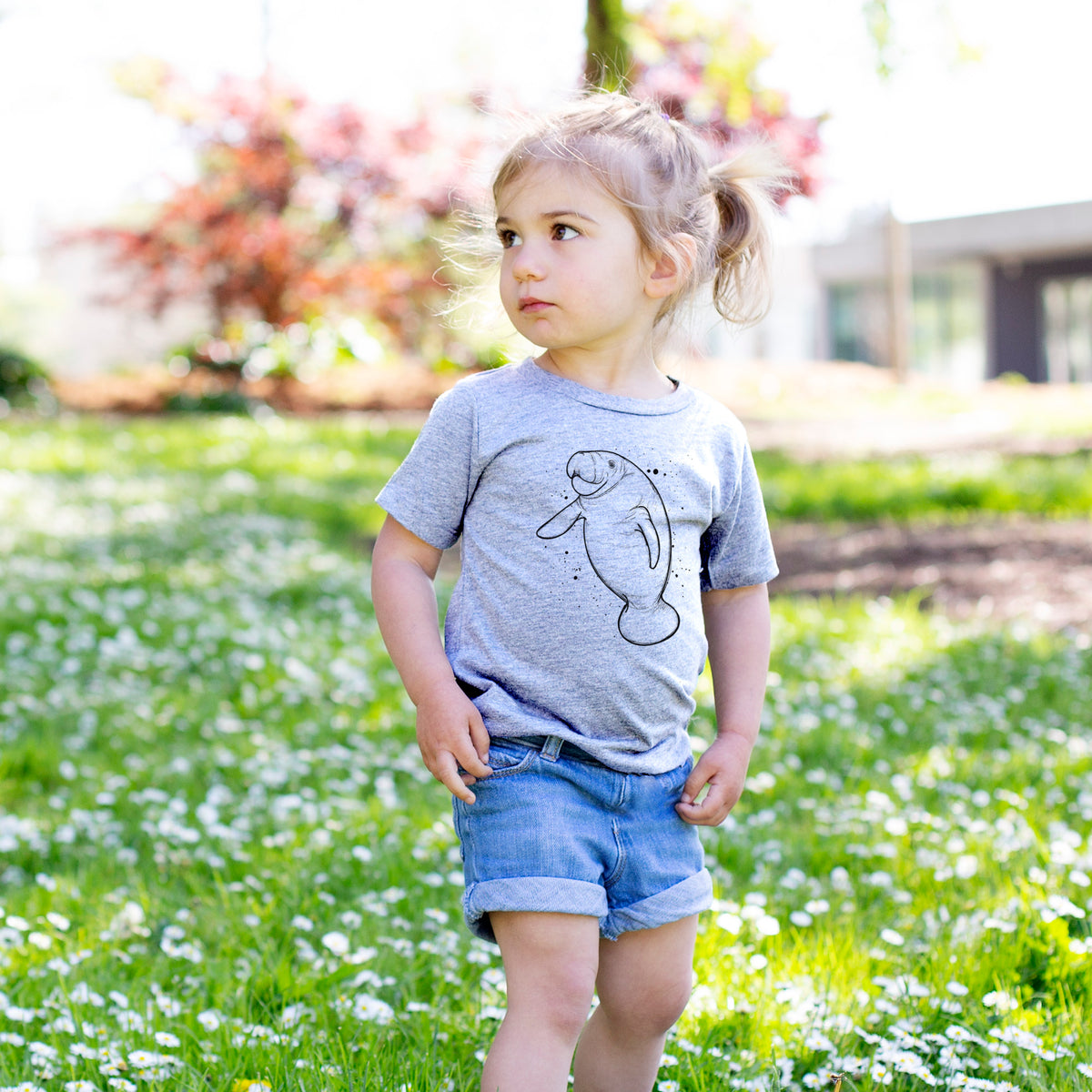 Toddler girl walking outdoors wearing a t-shirt printed with a black ink line drawing of a manatee swimming, with ink splatters.