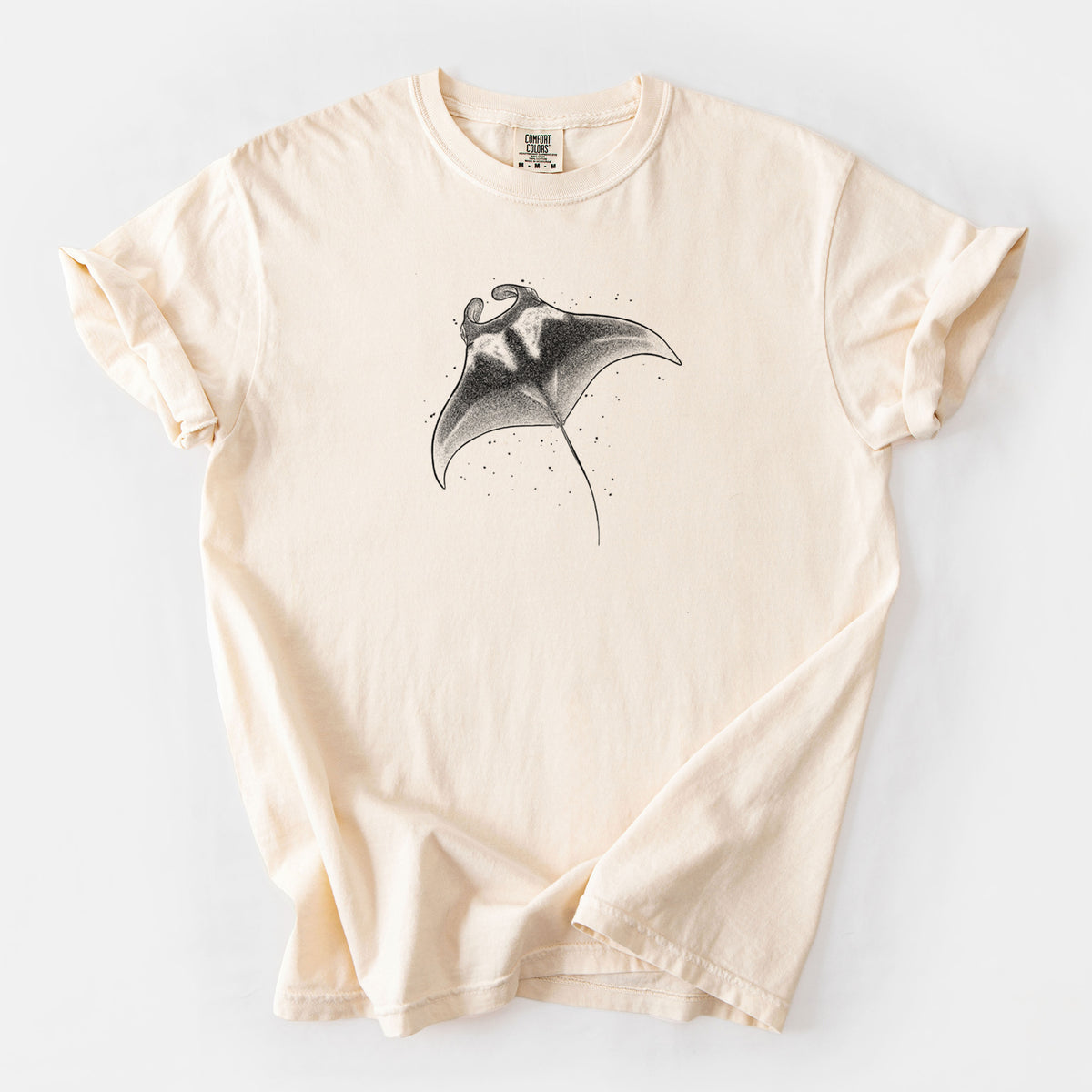 Ivory Comfort Colors t-shirt laid flat, printed with a black ink illustration of a manta ray gliding.