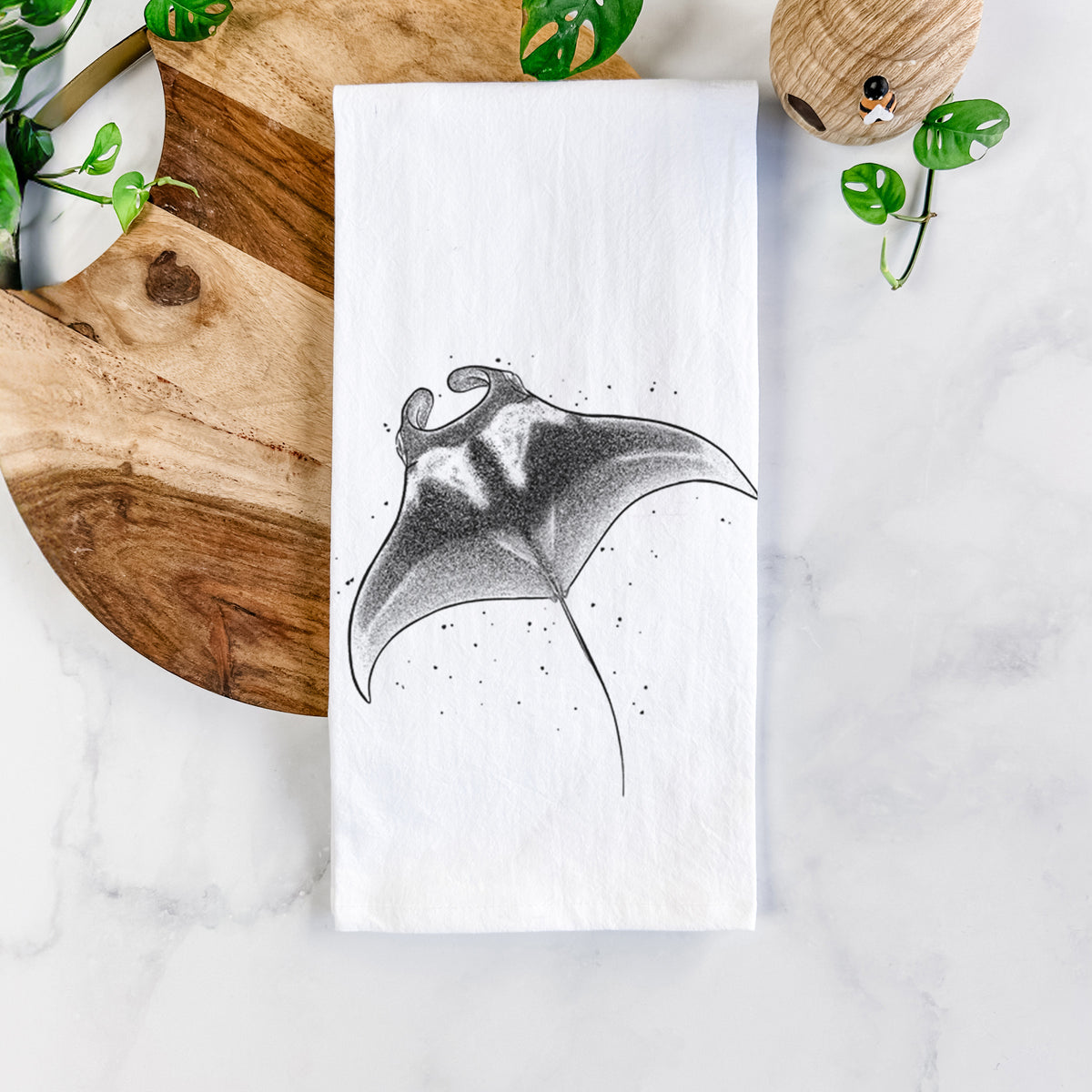 White cotton tea towel printed with a black ink illustration of a manta ray gliding, on a marble counter beside a round wooden cutting board and greenery.