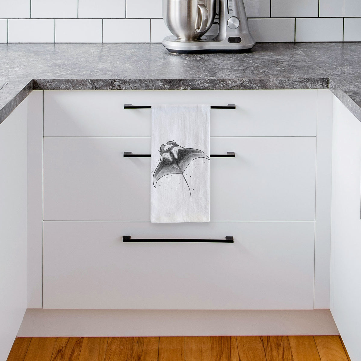White tea towel printed with a black ink illustration of a manta ray gliding, hanging from a drawer handle in a bright white kitchen.