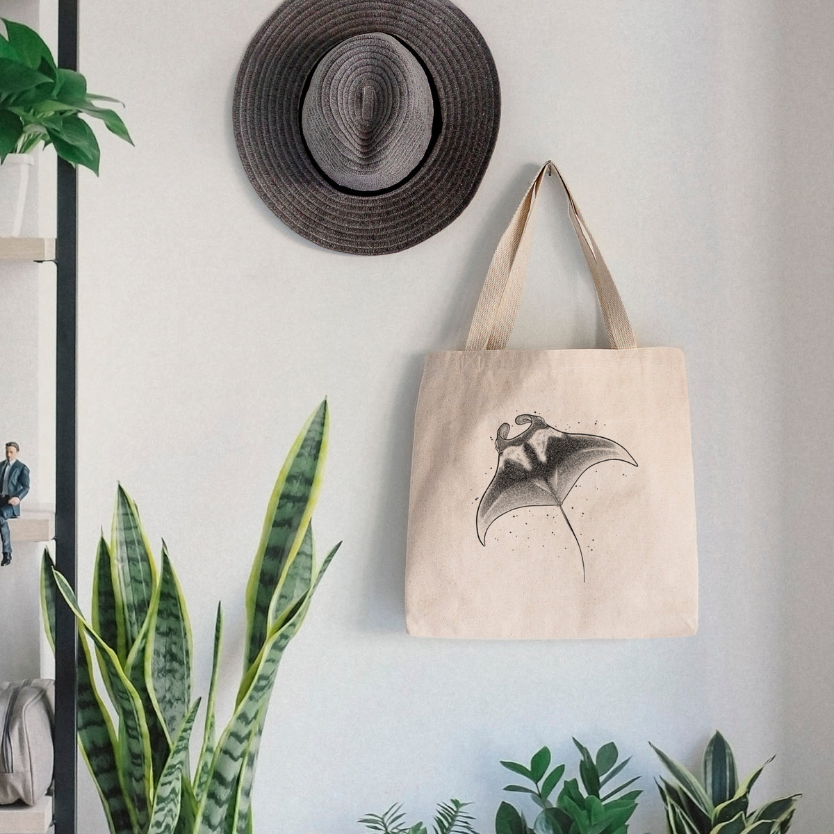 Canvas tote bag printed with a black ink illustration of a manta ray gliding, hanging on a wall hook below a hat, next to potted plants.