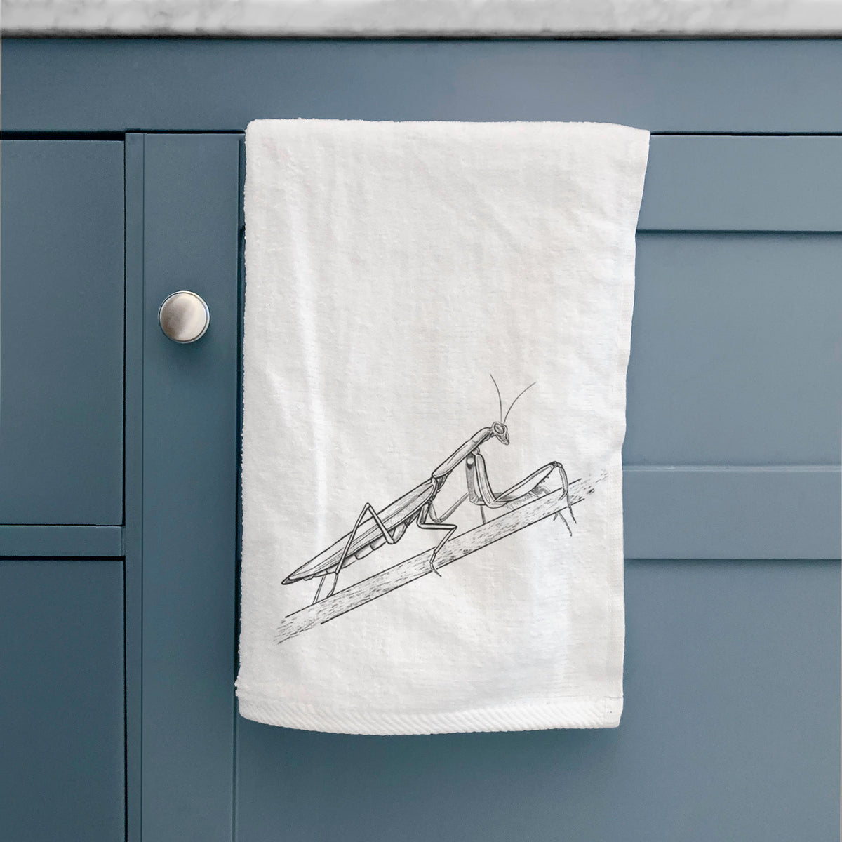 White hand towel printed with a detailed black ink illustration of a praying mantis on a stem, hanging on a cabinet door.