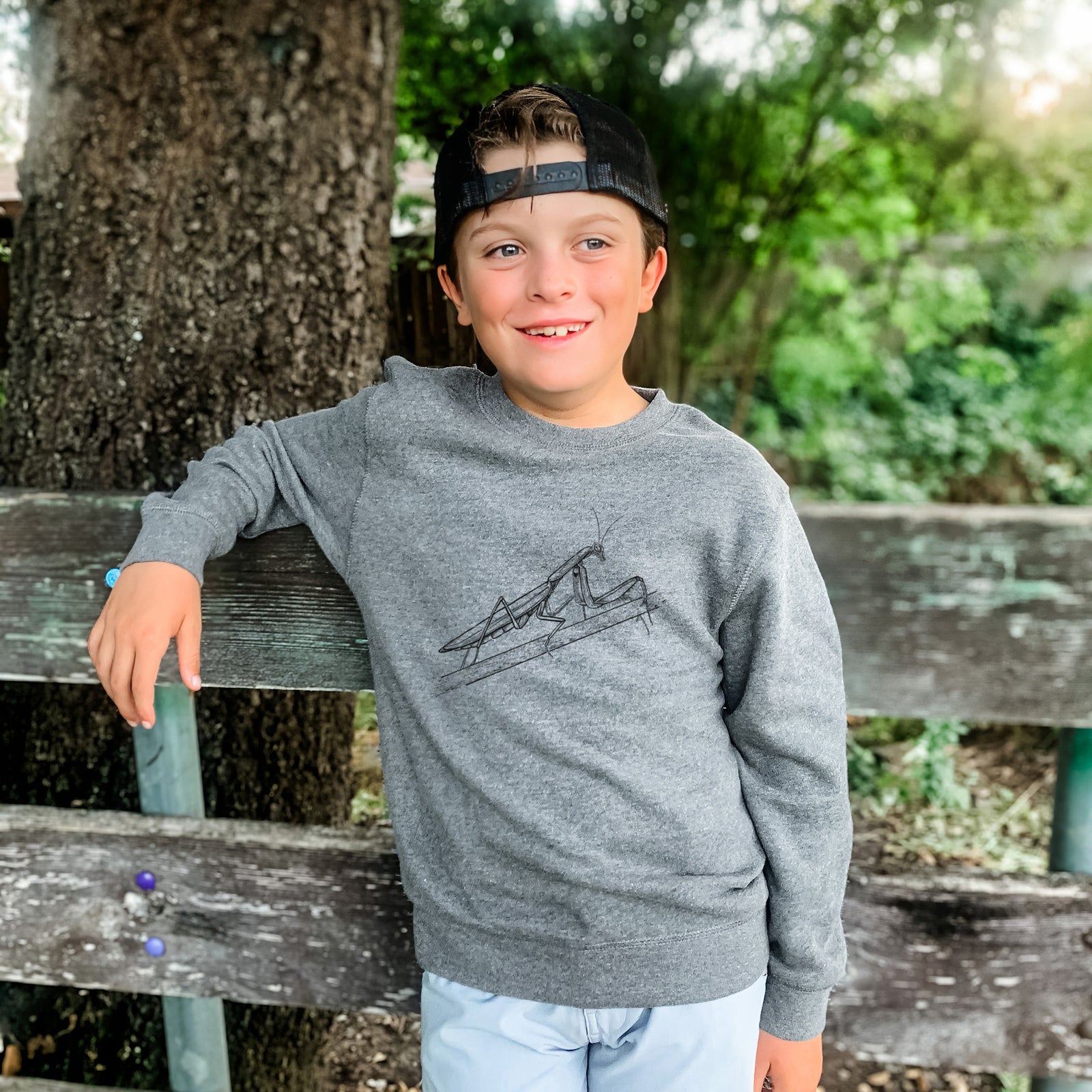 Boy wearing a youth crewneck sweatshirt printed with a detailed black ink illustration of a praying mantis on a stem, outdoors.