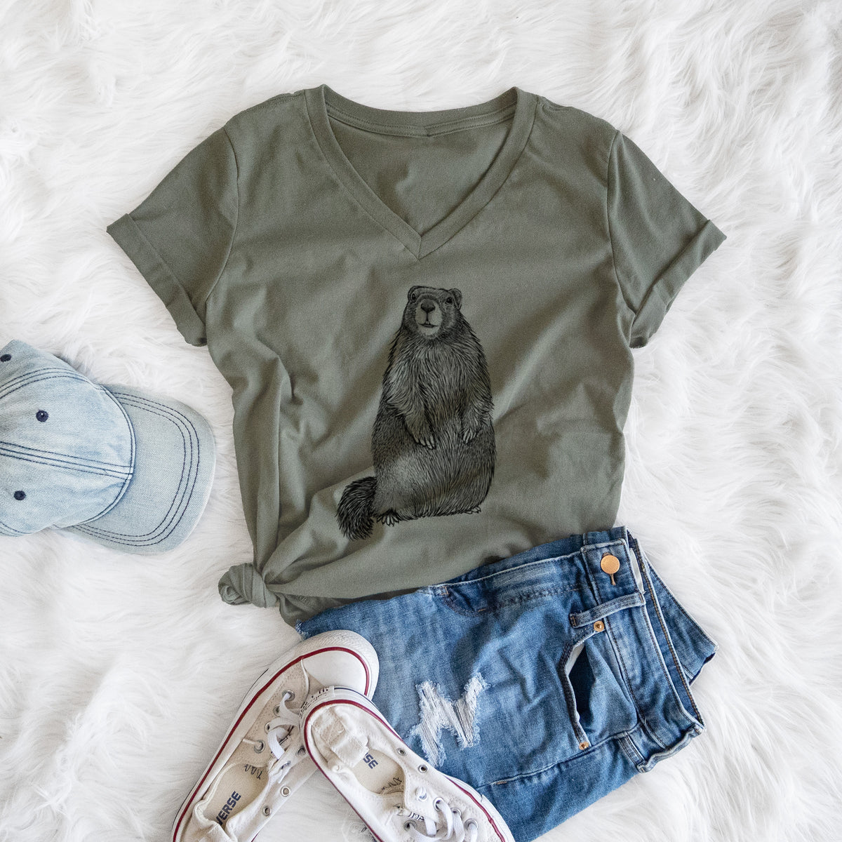 Military green women's relaxed fit cotton V-neck t-shirt printed with a detailed black ink illustration of a marmot standing upright, styled flat on a white faux fur rug with a cap, jeans and sneakers.