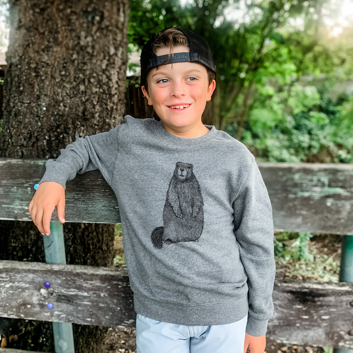 Boy wearing a youth crewneck sweatshirt printed with a detailed black ink illustration of a marmot standing upright, outdoors.