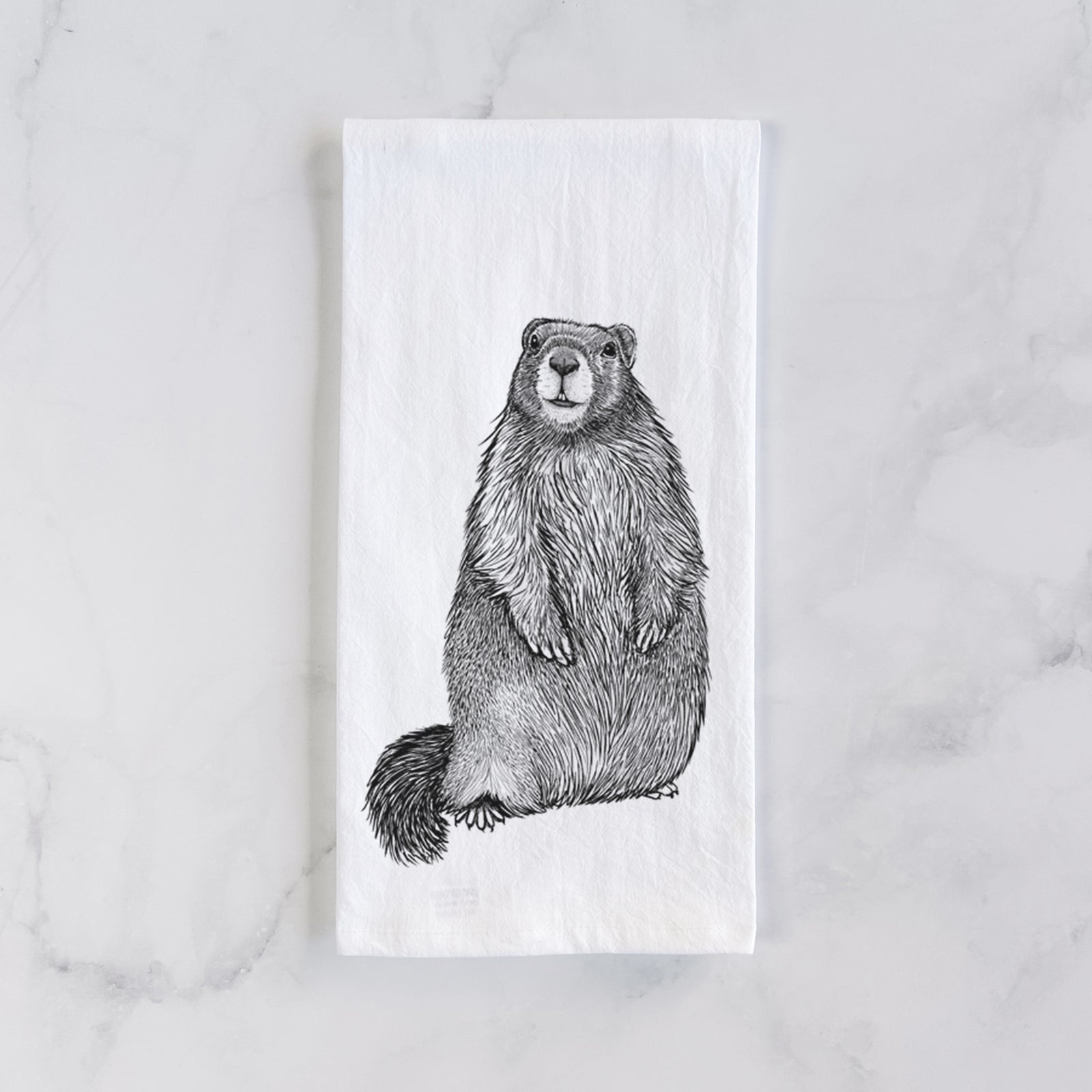 White cotton tea towel printed with a detailed black ink illustration of a marmot standing upright, on a marble counter beside a round wooden cutting board and greenery.