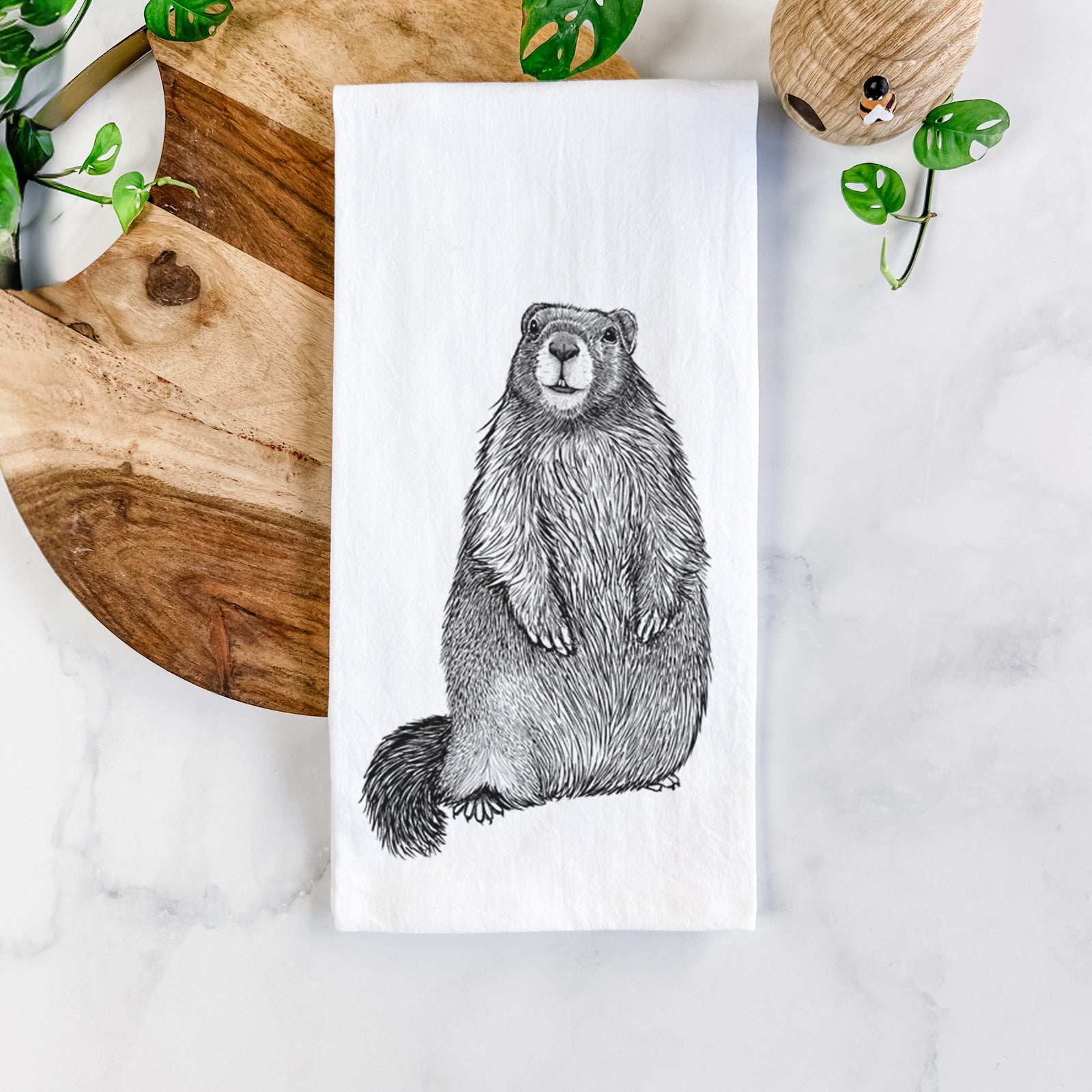 White cotton tea towel printed with a detailed black ink illustration of a marmot standing upright, on a marble counter beside a round wooden cutting board and greenery.