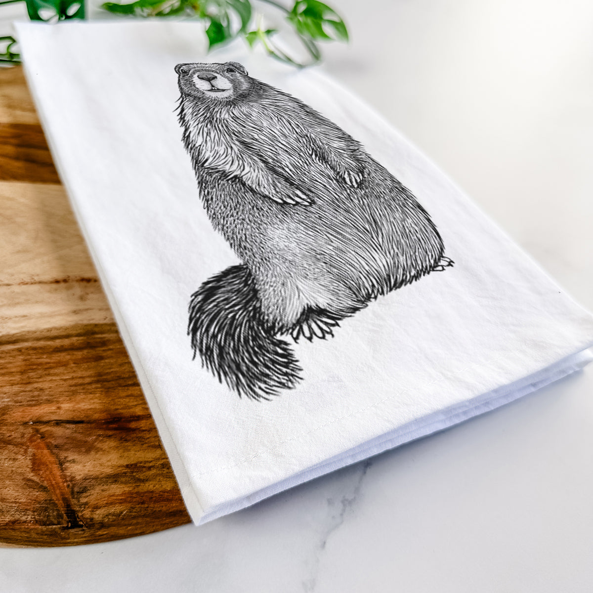 Close-up of a folded white tea towel on a wooden board, showing a detailed black ink illustration of a marmot standing upright.