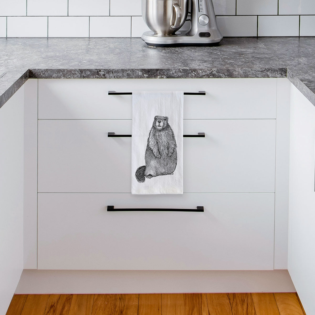 White tea towel printed with a detailed black ink illustration of a marmot standing upright, hanging from a drawer handle in a bright white kitchen.