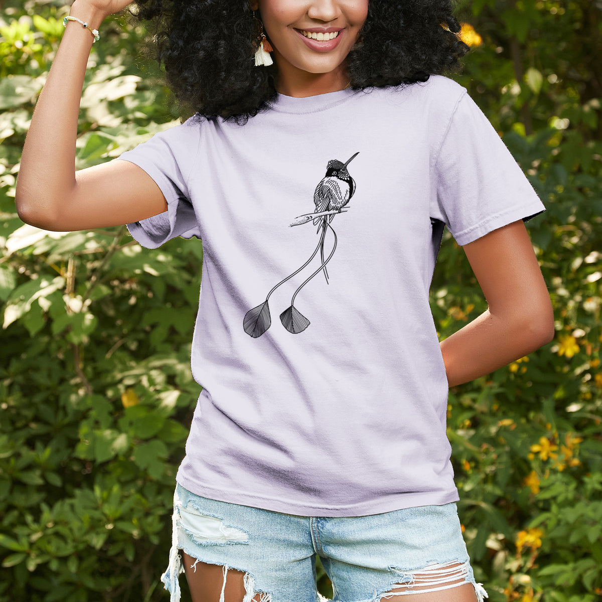 Woman wearing a Comfort Colors t-shirt printed with a black ink illustration of a marvelous spatuletail hummingbird perched, with its two long racket-tipped tail feathers, outdoors.