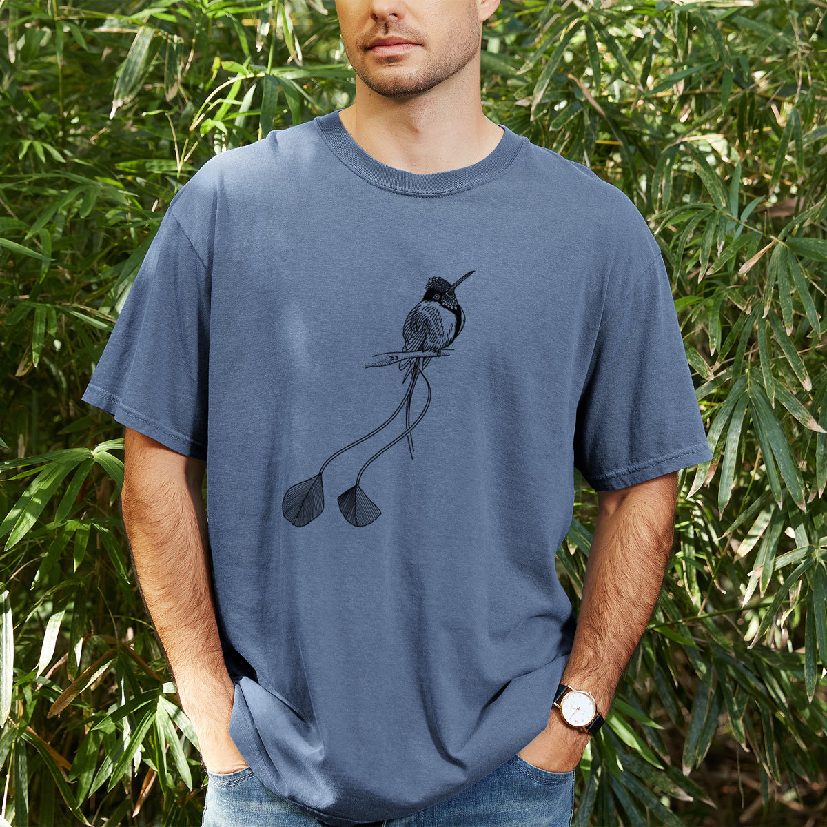 Man wearing a Comfort Colors t-shirt printed with a black ink illustration of a marvelous spatuletail hummingbird perched, with its two long racket-tipped tail feathers, outdoors.