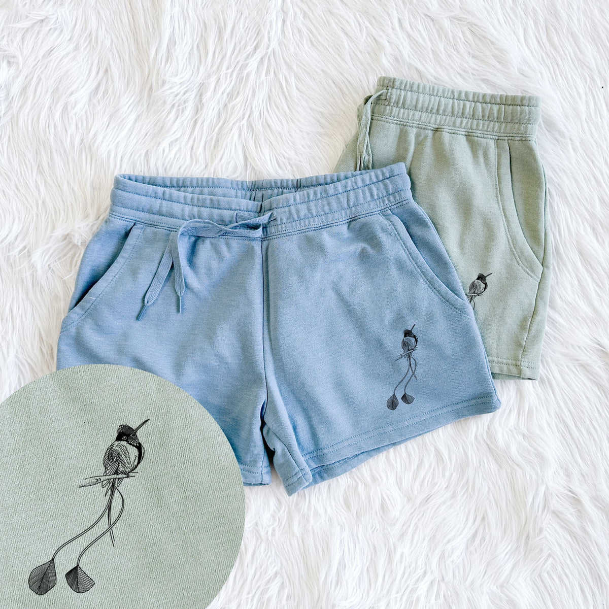 Two pairs of Cali Wave shorts in different colors printed with a black ink illustration of a marvelous spatuletail hummingbird perched, with its two long racket-tipped tail feathers, with a close-up of the print.