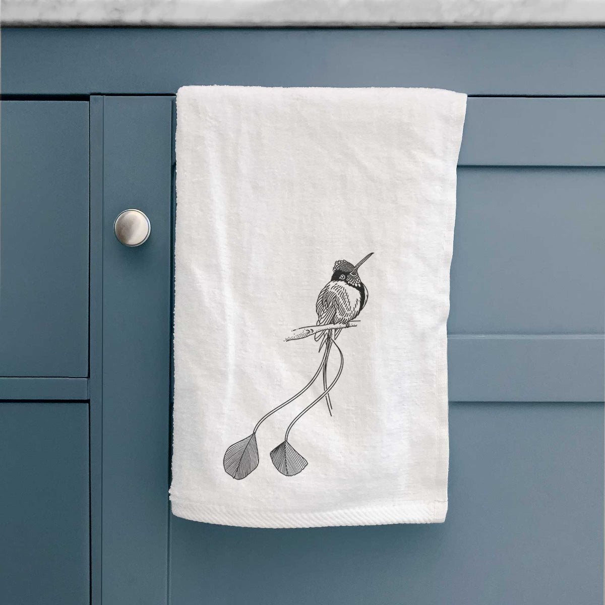White hand towel printed with a black ink illustration of a marvelous spatuletail hummingbird perched, with its two long racket-tipped tail feathers, hanging on a cabinet door.