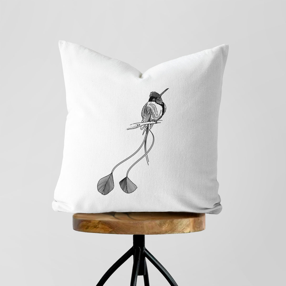 White pillow cover printed with a black ink illustration of a marvelous spatuletail hummingbird perched, with its two long racket-tipped tail feathers, on a wooden stool.