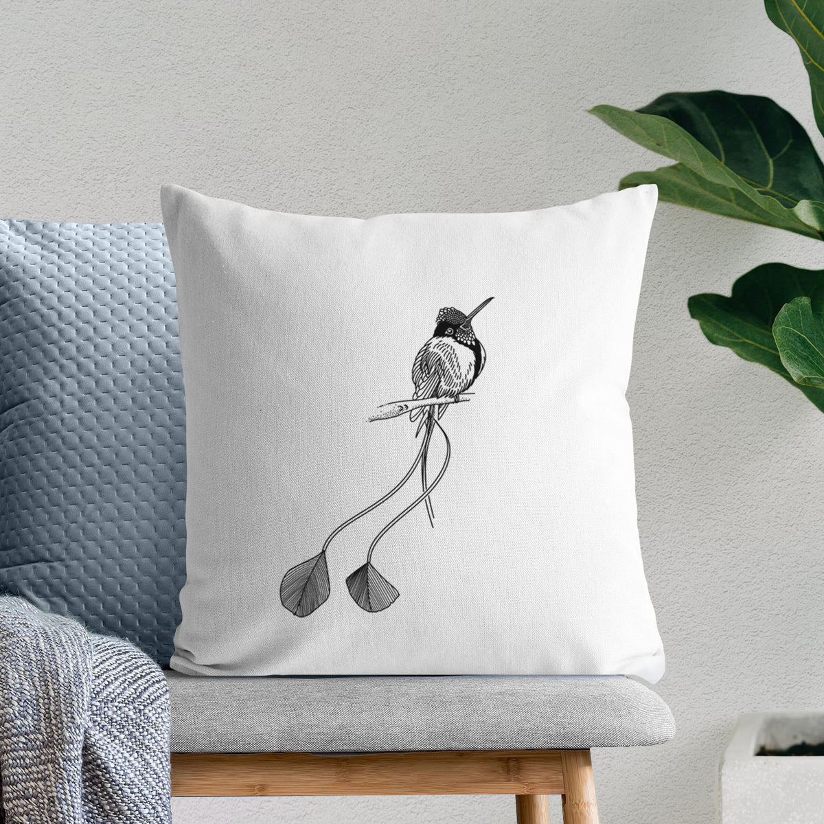 White pillow cover printed with a black ink illustration of a marvelous spatuletail hummingbird perched, with its two long racket-tipped tail feathers, on a bench beside plants.