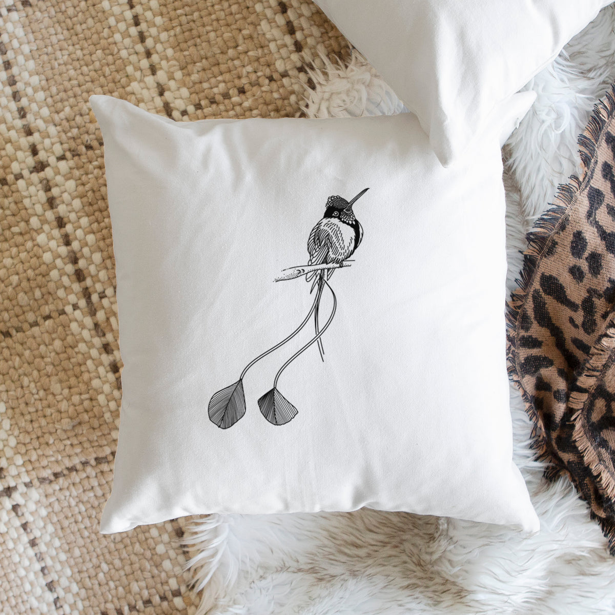 White pillow cover printed with a black ink illustration of a marvelous spatuletail hummingbird perched, with its two long racket-tipped tail feathers, on a woven chair.