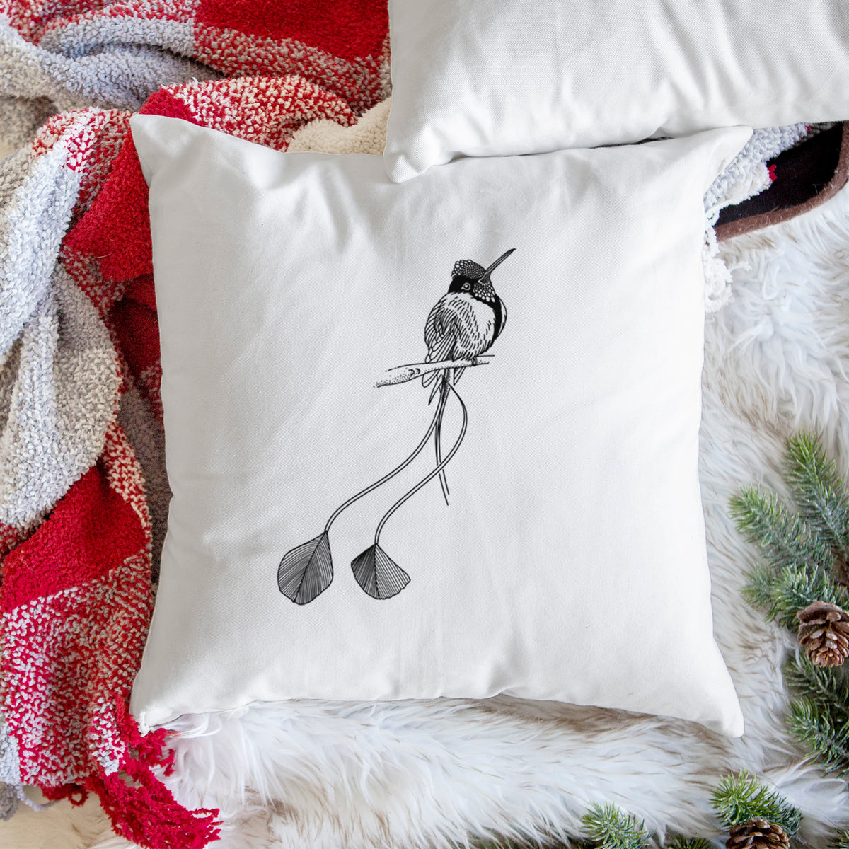 White pillow cover printed with a black ink illustration of a marvelous spatuletail hummingbird perched, with its two long racket-tipped tail feathers, on a woven chair.
