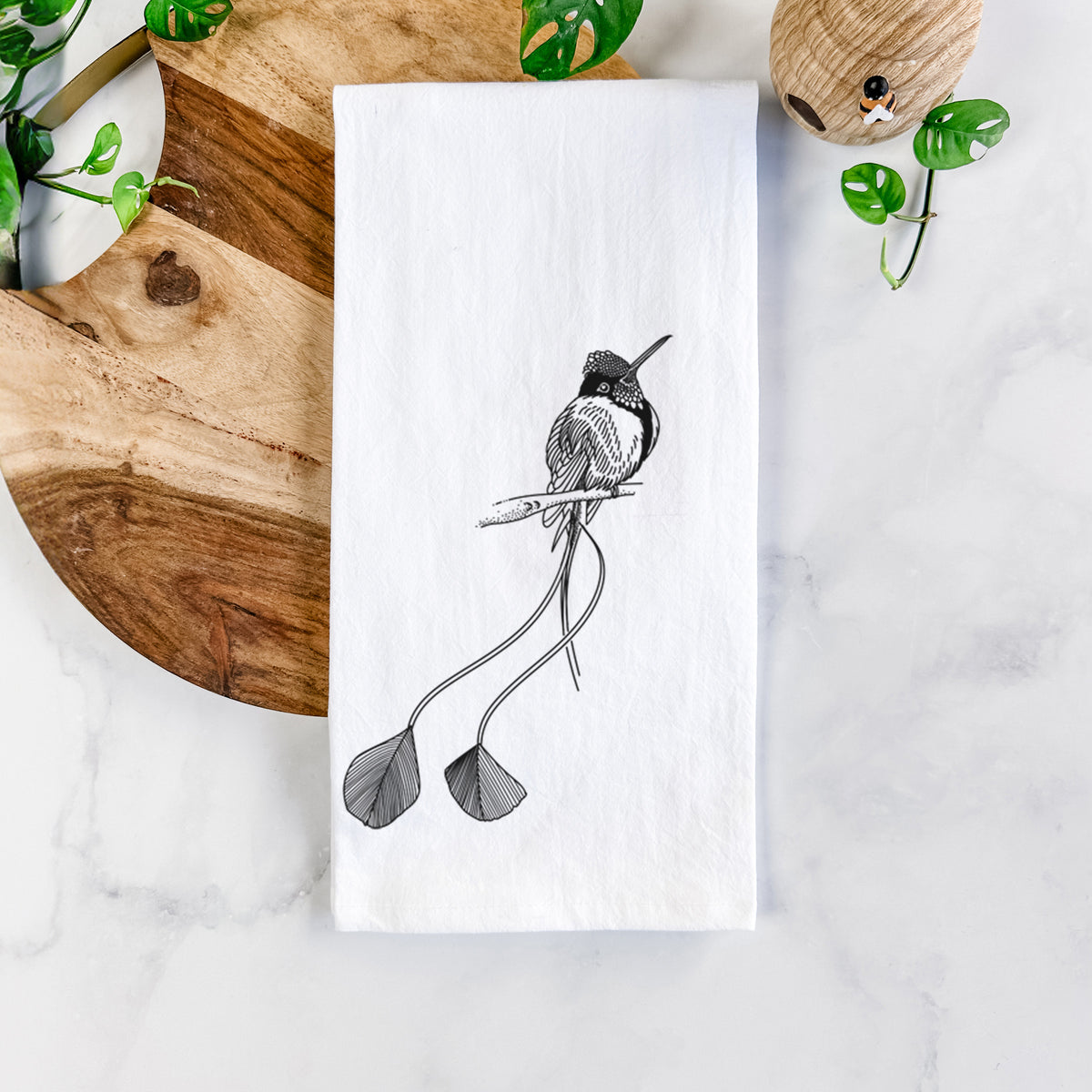 White cotton tea towel printed with a black ink illustration of a marvelous spatuletail hummingbird perched, with its two long racket-tipped tail feathers, on a marble counter beside a round wooden cutting board and greenery.