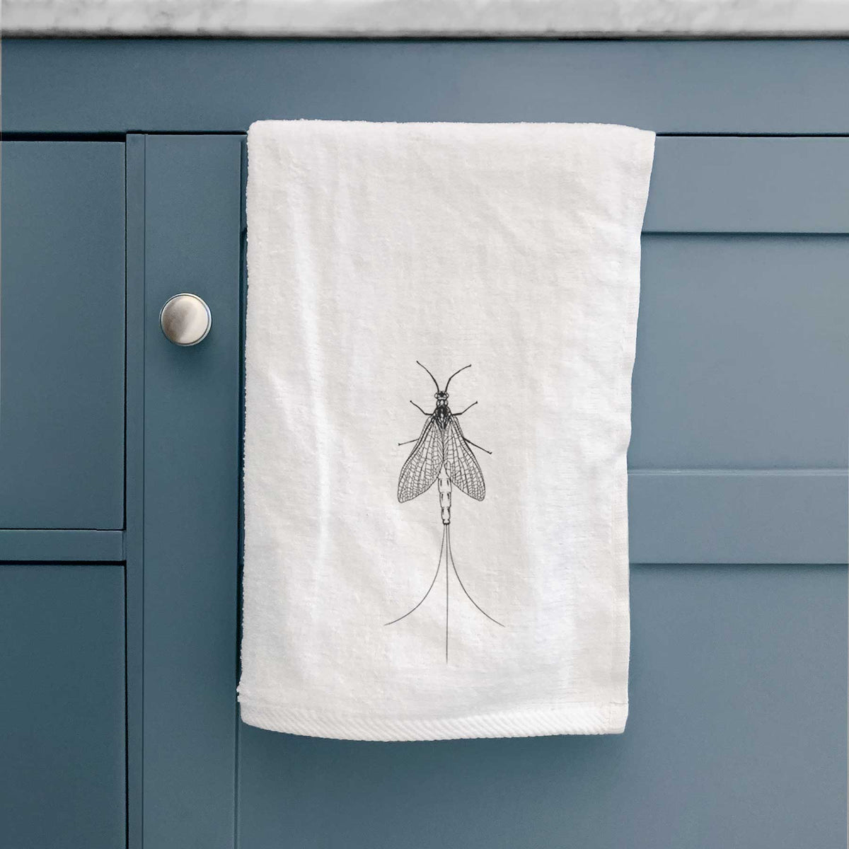 White hand towel printed with a detailed black ink illustration of a mayfly with its wings raised and long tail filaments, hanging on a cabinet door.