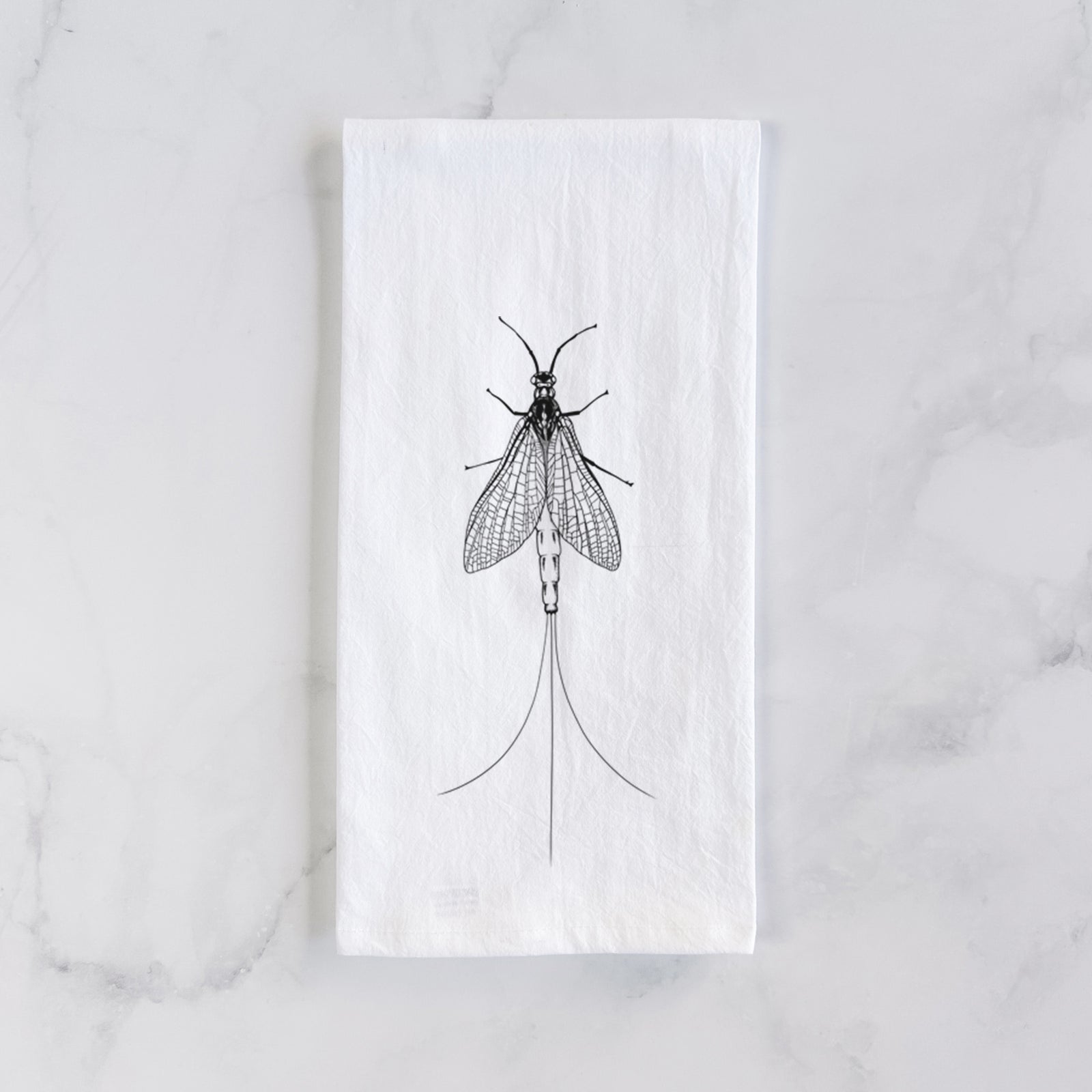 White cotton tea towel printed with a detailed black ink illustration of a mayfly with its wings raised and long tail filaments, on a marble counter beside a round wooden cutting board and greenery.