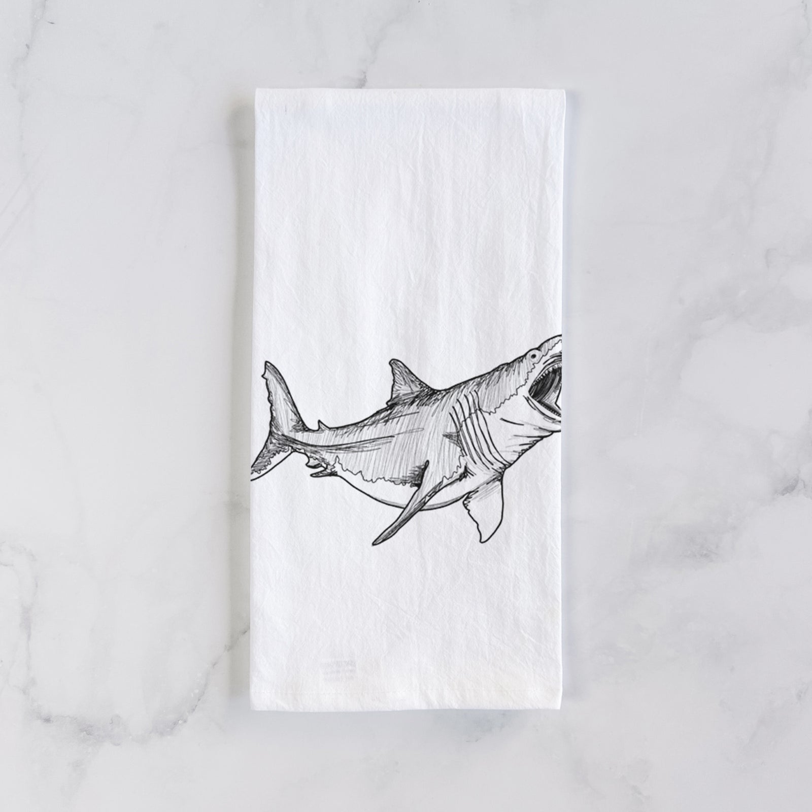 White cotton tea towel printed with a black ink illustration of a megalodon shark with its jaws open, on a marble counter beside a round wooden cutting board and greenery.