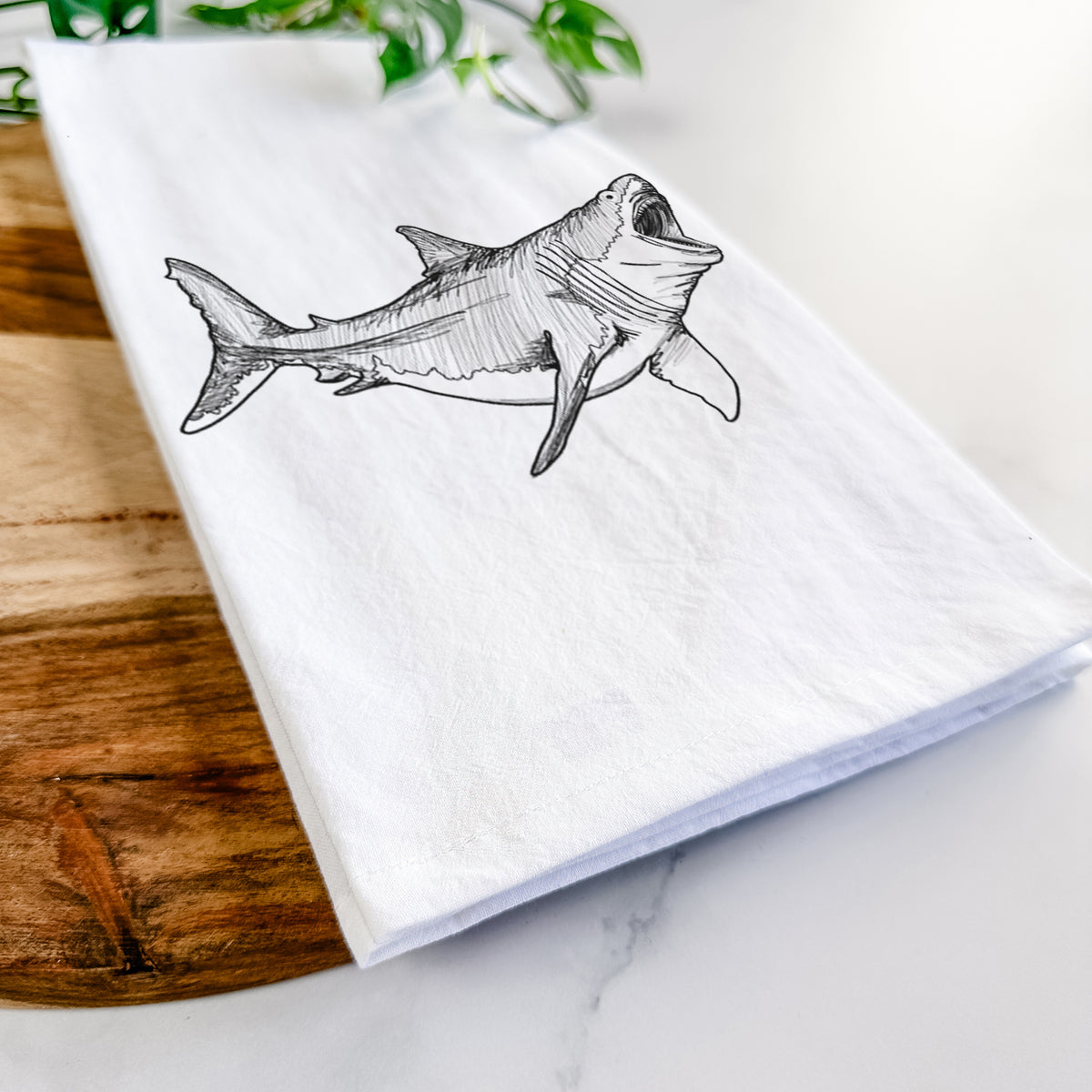 Close-up of a folded white tea towel on a wooden board, showing a black ink illustration of a megalodon shark with its jaws open.