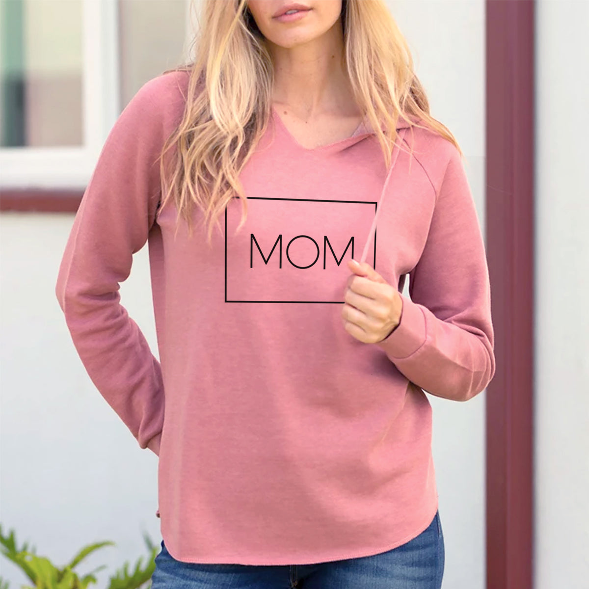 Smiling woman on a seaside bluff wearing a dusty rose women's Cali Wave hooded sweatshirt printed with the word MOM in thin black letters inside a rectangle outline.