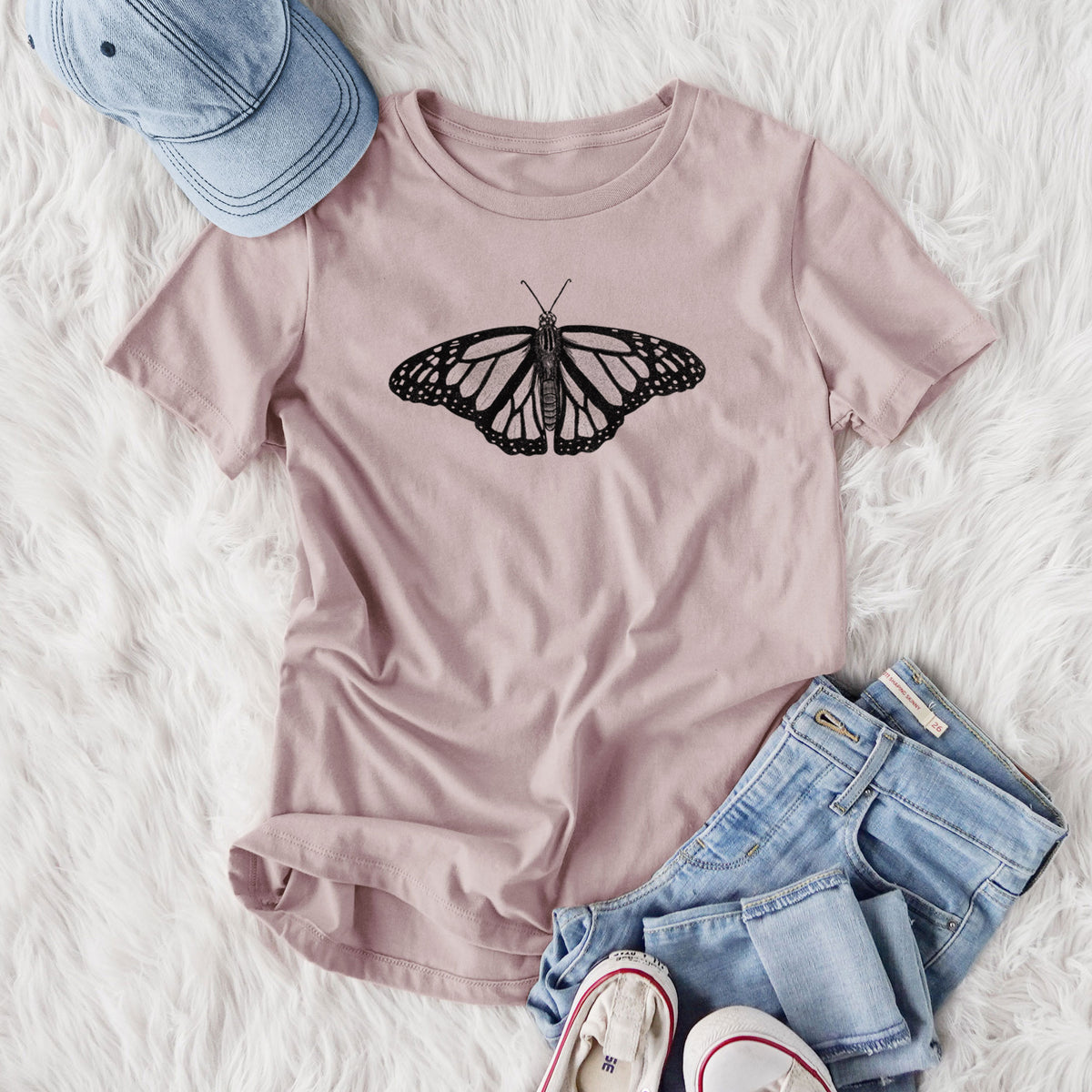 Women's relaxed fit cotton t-shirt printed with a detailed black ink illustration of a monarch butterfly with its wings spread, styled flat on a white faux fur rug with jeans.