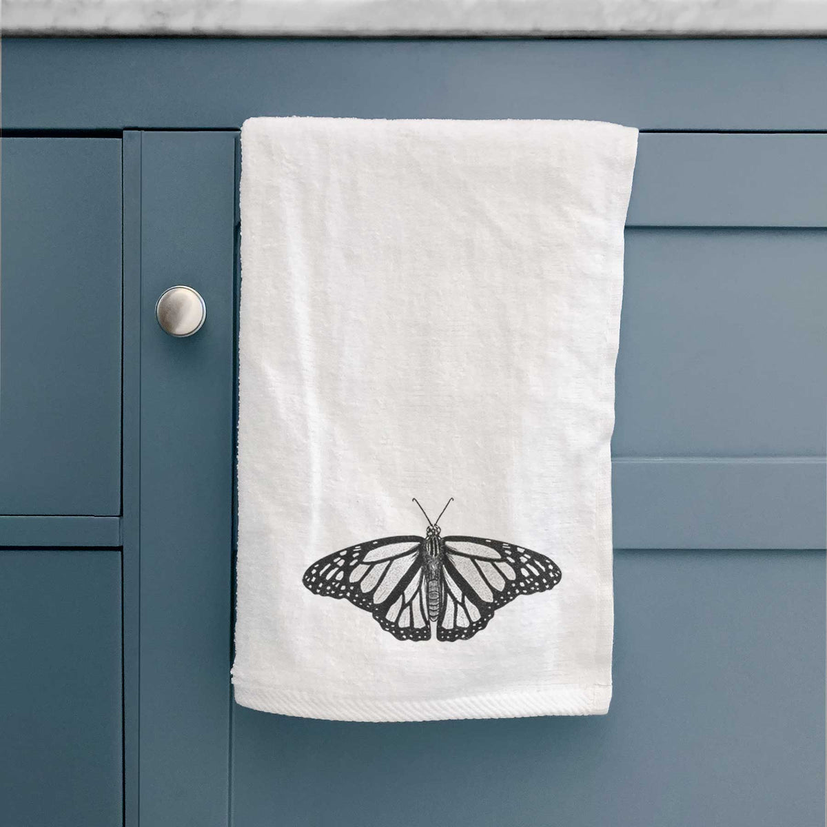 White hand towel printed with a detailed black ink illustration of a monarch butterfly with its wings spread, hanging on a cabinet door.