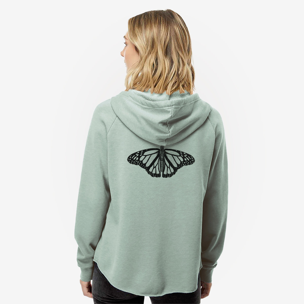 Woman wearing a sage green women's Cali Wave zip-up sweatshirt printed with a detailed black ink illustration of a monarch butterfly with its wings spread, seen from behind.