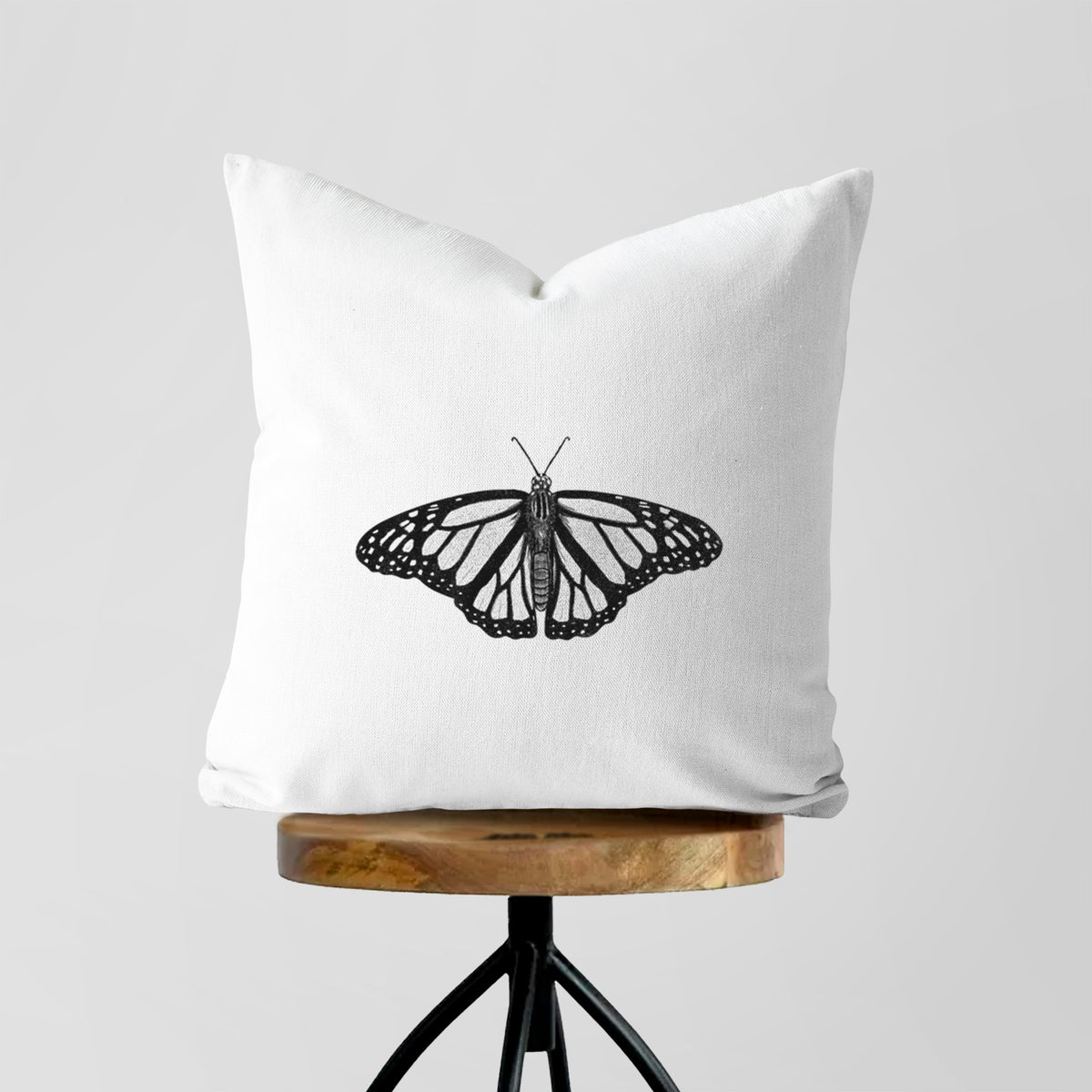 White pillow cover printed with a detailed black ink illustration of a monarch butterfly with its wings spread, on a wooden stool.