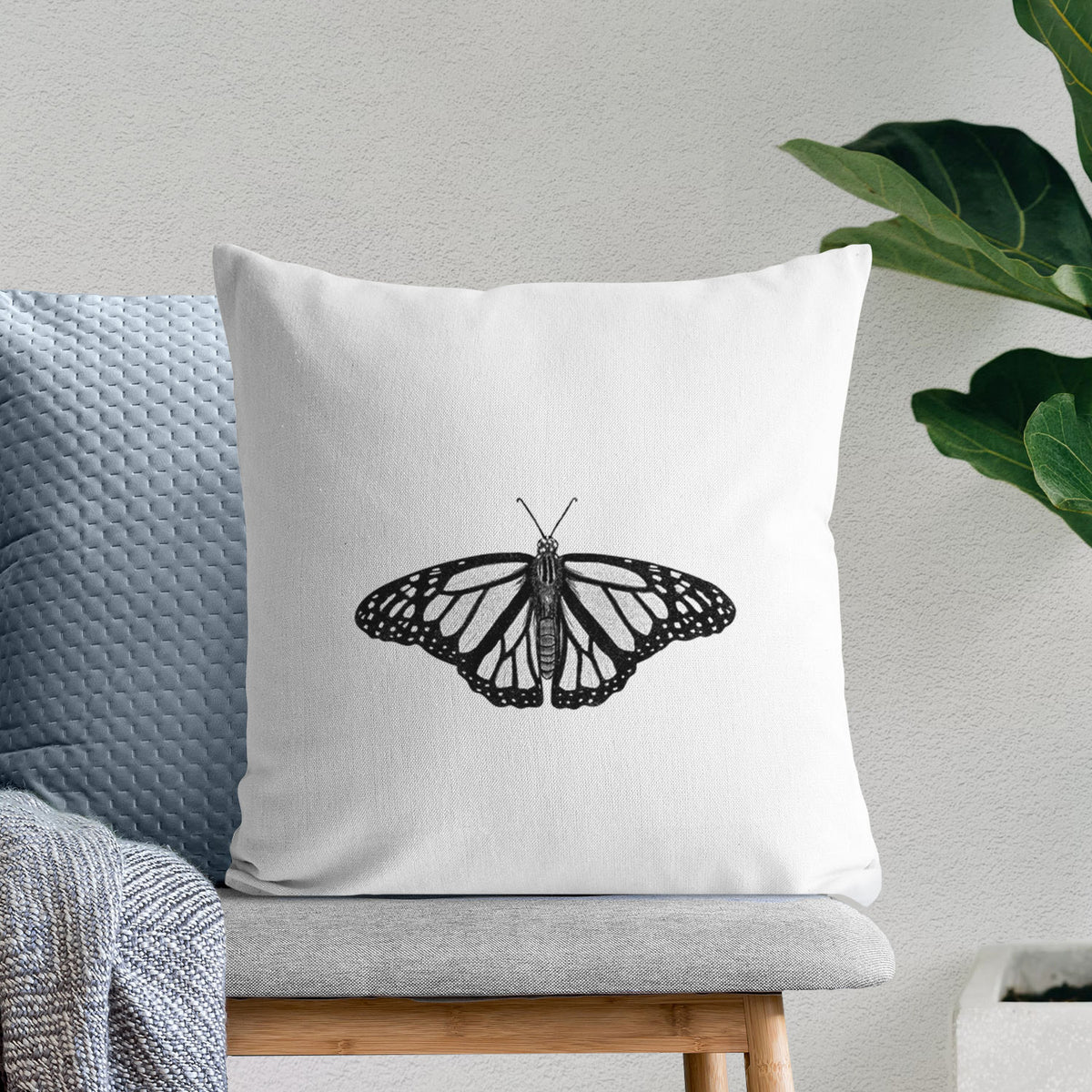 White pillow cover printed with a detailed black ink illustration of a monarch butterfly with its wings spread, on a bench beside plants.