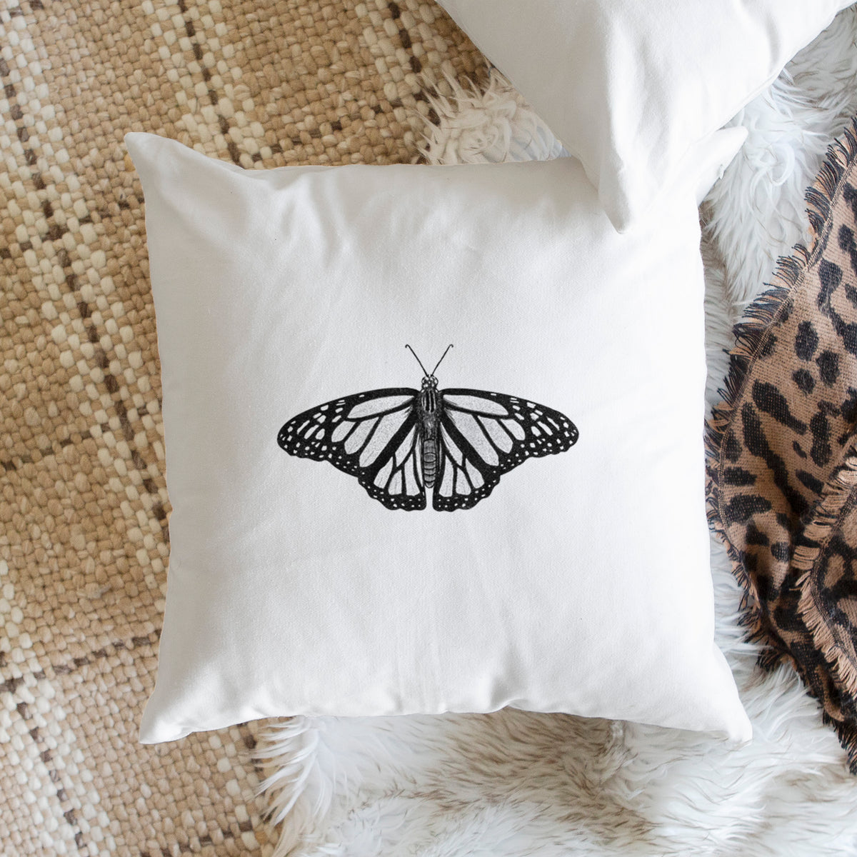 White pillow cover printed with a detailed black ink illustration of a monarch butterfly with its wings spread, on a woven chair.
