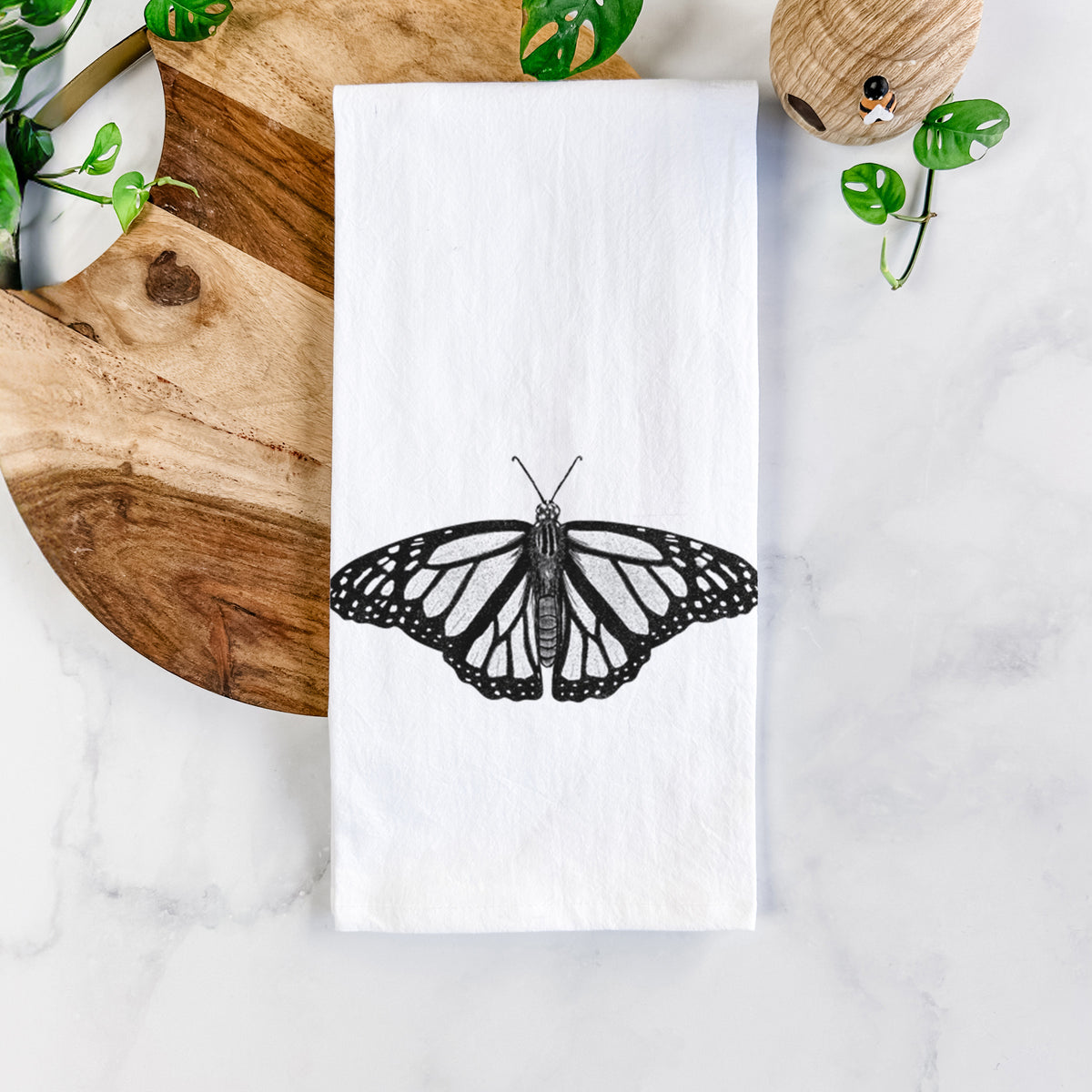 White cotton tea towel printed with a detailed black ink illustration of a monarch butterfly with its wings spread, on a marble counter beside a round wooden cutting board and greenery.